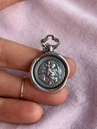 A hand holds a St Christopher Charm by Jessica de Lotz Jewellery, featuring a raised seated figure with outstretched arms and an animal on oxidised silver, resting on light pink fabric.