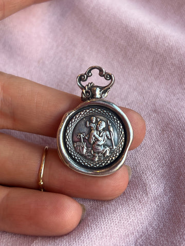 A hand holds a St Christopher Charm by Jessica de Lotz Jewellery, featuring a raised seated figure with outstretched arms and an animal on oxidised silver, resting on light pink fabric.