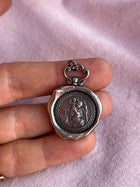 A hand holds a round oxidised silver St Christopher Charm by Jessica de Lotz Jewellery, featuring a raised scene with rays and floral details. The pendant rests on soft pink fabric.
