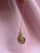 The St Christopher Charms necklace by Jessica de Lotz Jewellery features a round, textured gold pendant with an engraved figure, inspired by vintage designs, suspended from a fine chain and displayed on soft pink fabric.