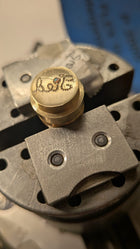 A close-up of the Tied Charm by Jessica de Lotz Jewellery shows a round brass wax seal secured in a vise, featuring cursive engraving and an inlaid colored stone—perfect as personalized friendship jewelry. Blue and metallic tones blur in the background.