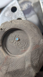 A small opal gemstone sits in the center of a circular indentation on a textured surface. Reminiscent of a wax seal charm, the Tied Charm by Jessica de Lotz Jewellery is seen with metal tools blurred in the background.