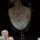 A woman in a white lace camisole is seen reflected in an oval mirror, several pendant necklaces draped around her neck—including the Practical Magic Chatelaine Hand Necklace (Charm & Chain Bundle) by Jessica de Lotz Jewellery. The scene is softly illuminated by the warm light of three white candles burning in the foreground, their glow highlighting her collection of Practical Magic charms. Her face is partially out of the frame.