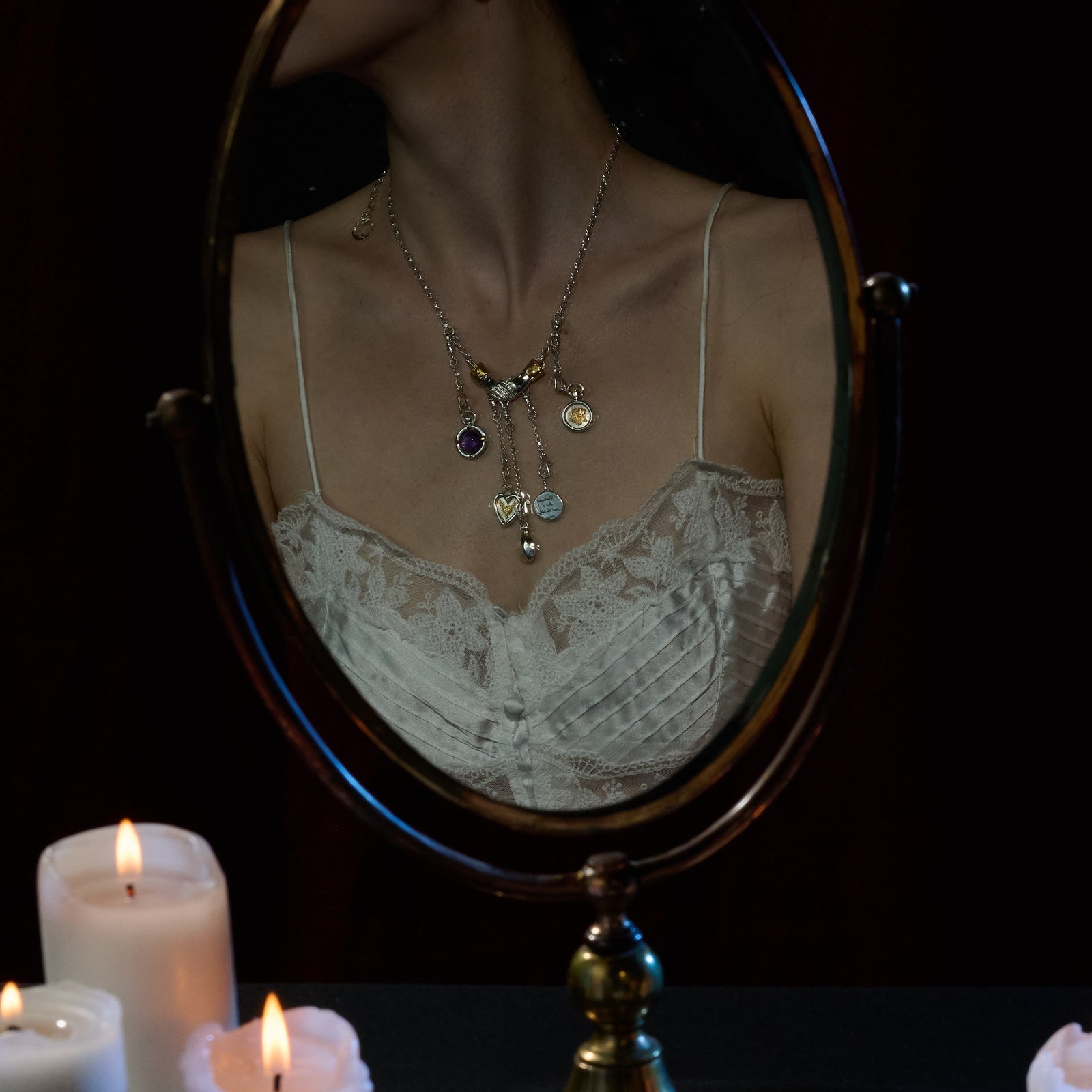 A woman in a white lace camisole is seen reflected in an oval mirror, several pendant necklaces draped around her neck—including the Practical Magic Chatelaine Hand Necklace (Charm & Chain Bundle) by Jessica de Lotz Jewellery. The scene is softly illuminated by the warm light of three white candles burning in the foreground, their glow highlighting her collection of Practical Magic charms. Her face is partially out of the frame.