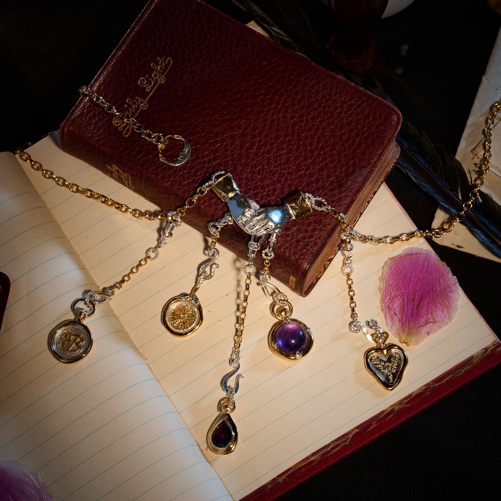 The Practical Magic Chatelaine Hand Necklace (Charm & Chain Bundle) by Jessica de Lotz Jewellery, featuring five gold and silver pendants, is elegantly draped across an open lined notebook, a closed maroon Holy Bible, and scattered pink rose petals. Nearby rests a dark feather pen, completing the vintage-inspired scene.