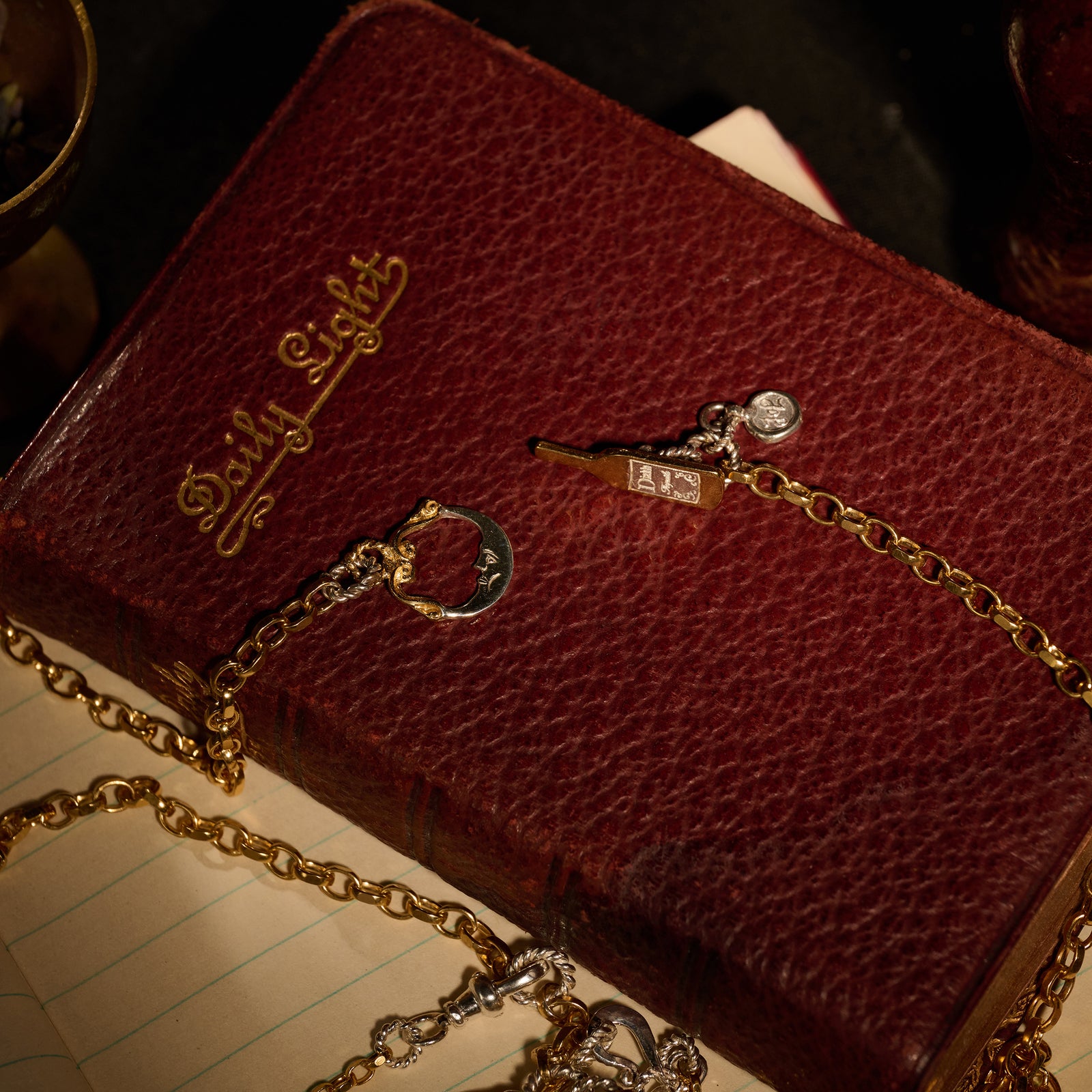 A red leather Daily Light book rests atop a lined notepad. Two gold chains from Jessica de Lotz Jewellery’s Practical Magic Chatelaine Hand Necklace (Charm & Chain Bundle) are draped over the book: one features a crescent-shaped charm reminiscent of Practical Magic, and the other displays a delicate bar and circular pendant. A metal cup peeks in from the corner.