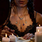 A woman in a black lace top sits at a candlelit table, her silver Practical Magic Chatelaine Hand Necklace from Jessica de Lotz Jewellery catching the glow as she looks down, partially obscured by shadows. She holds a wax seal stamp and appears to be sealing a letter amidst the flickering candlelight.