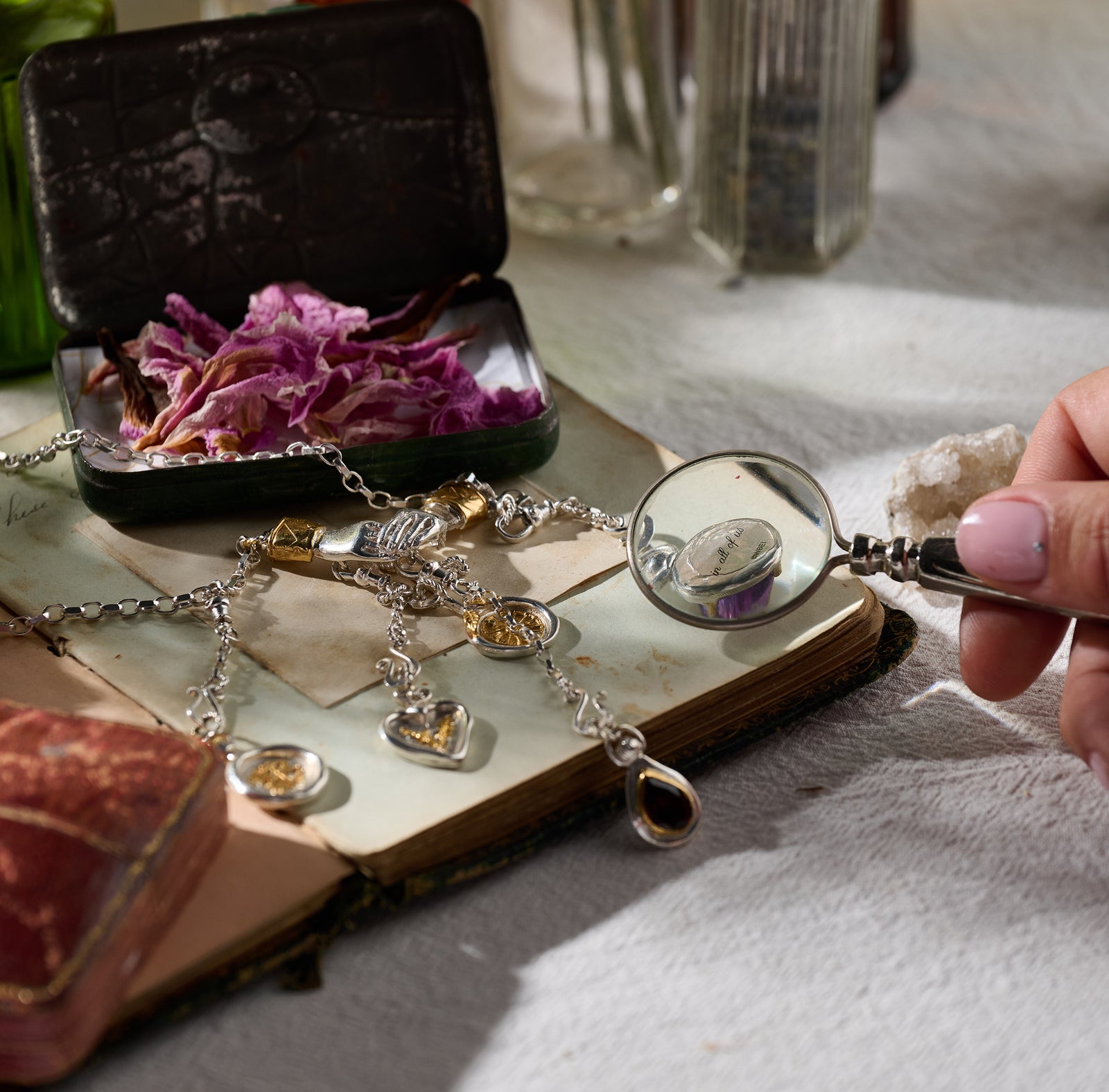 A hand holds a magnifying glass over a Jessica de Lotz Jewellery Practical Magic Chatelaine Hand Necklace (Charm & Chain Bundle), which rests atop an old open book. Nearby lie a worn red book, dried purple flowers, a green glass bottle, and softly blurred bottles in the background. Sunlight filters in, casting gentle shadows across the scene.