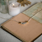 The Practical Magic Chatelaine Hand Necklace (Charm & Chain Bundle) by Jessica de Lotz Jewellery, featuring a silver handshake pendant with gold accents, rests atop the pages of an open vintage book beside a softly glowing white candle. Set on a light fabric surface and bathed in gentle natural light, the scene captures a cozy, whimsical atmosphere reminiscent of magical charms and the distinctive artistry characteristic of Jessica de Lotz Jewellery.