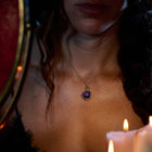 A woman wearing a black lace top and the Jessica de Lotz Jewellery ’There’s A Little Witch In All Of Us’ Charm necklace, featuring a purple gemstone pendant, sits near burning white candles. Her face is partially visible in the warm light, and her wavy hair cascades over her shoulders. A red object appears at the left edge of the scene.