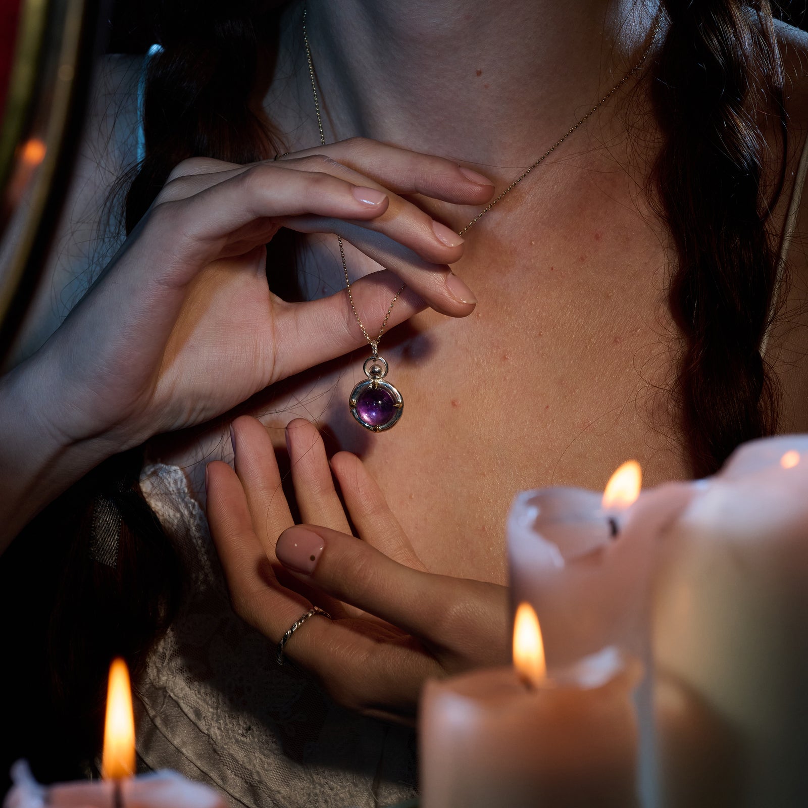 A woman with braided hair holds the “There’s A Little Witch In All Of Us Charm” necklace by Jessica de Lotz Jewellery—a round, purple pendant reminiscent of a chatelaine—close to her chest. Her nails are manicured and she wears a white top, while softly glowing candles with melted wax in the foreground cast a warm light over her hands and chest.