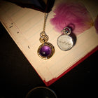 A gold hand necklace featuring a purple gemstone pendant, together with Jessica de Lotz Jewellery’s “There’s A Little Witch In All Of Us” round silver charm, rests atop a lined notebook. Pink flower petals and delicate wax seal charms are scattered nearby, while the edge of a magnifying glass peeks into the scene. The warm, dramatic lighting evokes the enchanting atmosphere of Practical Magic 2.