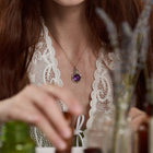A woman with long brown hair, dressed in a lace white top, sits at a table handling small bottles. Around her neck shines Jessica de Lotz Jewellery’s “There’s A Little Witch In All Of Us” charm, its silver chatelaine design finished with a purple pendant. Blurred jars and sprigs of lavender in the foreground evoke the enchanting spirit of Practical Magic jewelry.