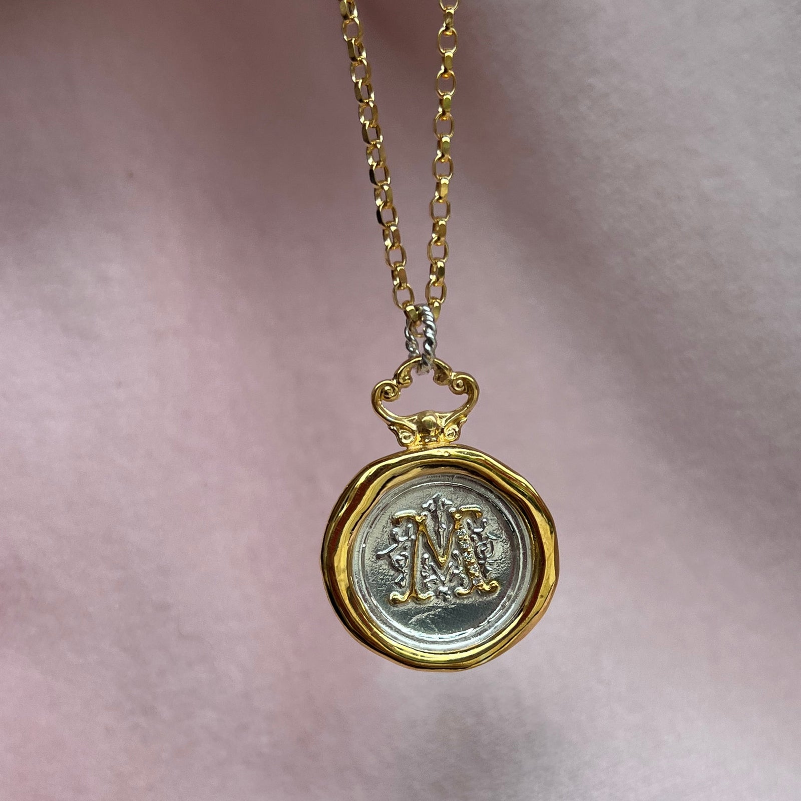 The Stone Selection Add-On from JdL Jewellery lets you set stones for extra shine, adding sparkle to your piece. Shown here on a gold chain with a round, two-tone pendant and ornate center “M”, beautifully displayed against soft pale pink fabric.