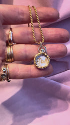 A hand adorned with gold rings holds Jessica de Lotz Jewellery's Tied Charm—a gold chain necklace with a round, wax seal-style pendant engraved with "TIED" on white—resting against pale pink fabric.