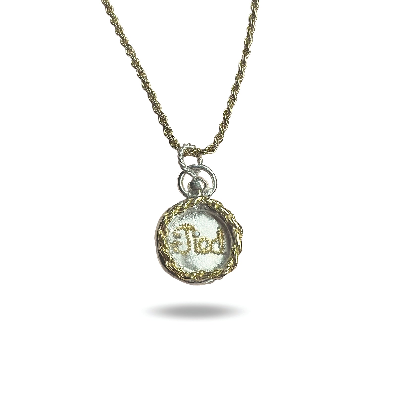 The Tied Charm necklace by Jessica de Lotz Jewellery features a gold rope chain and round pendant edged in twisted gold and silver. Inside, white fabric is embroidered with “Fidel” in gold thread, all casting a soft shadow on a white background.
