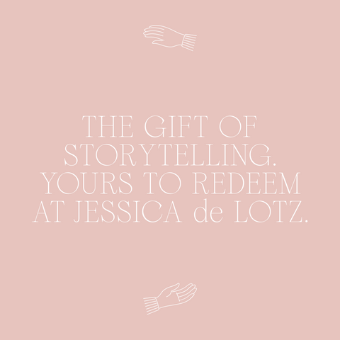 Centered white text on a pale pink background reads, THE GIFT OF STORYTELLING. REDEEM YOUR JESSICA DE LOTZ GIFT CARD AT JDL JEWELLERY. Simple white line drawings of open hands frame the text above and below.