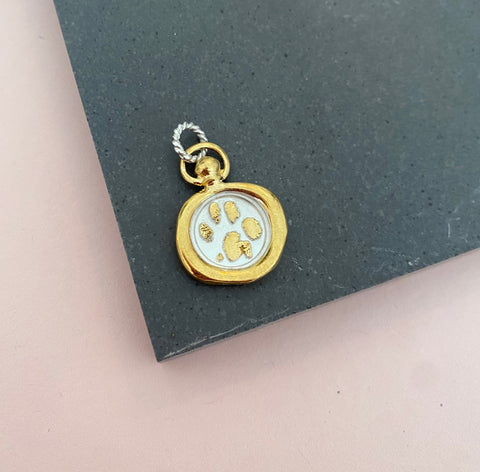 The Hand or Paw Imprint Charm by Jessica de Lotz Jewellery features a round pendant with a raised gold rim and white center showcasing a gold paw print, reminiscent of a wax seal, displayed on a dark gray surface with a hint of soft pink background.